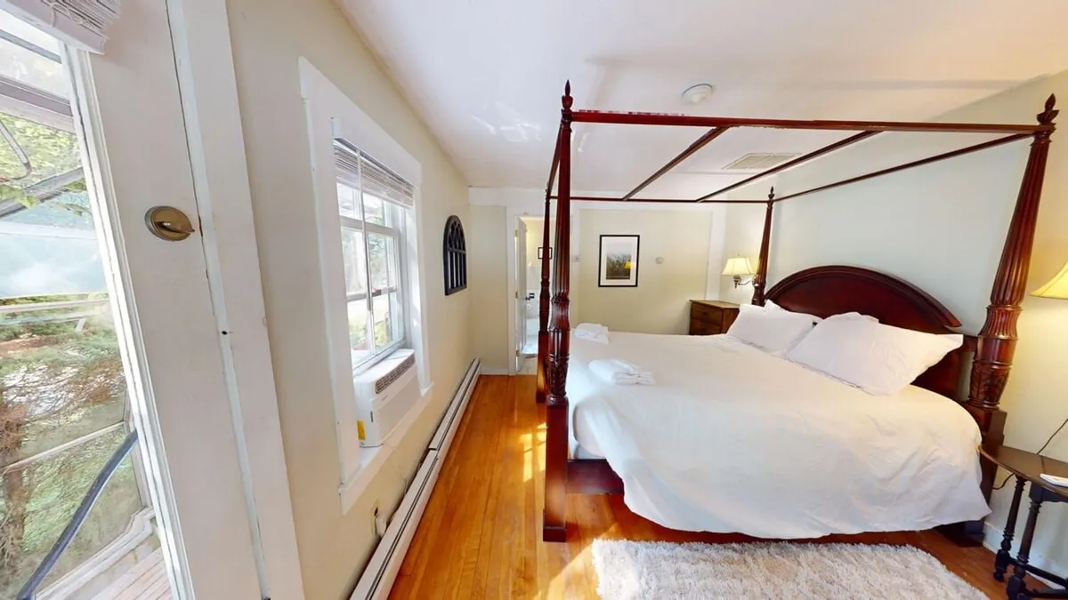 A serene bedroom featuring a four-poster bed draped in soft white linens, large windows letting in abundant natural light, and warm wooden floors. The tranquil environment is enhanced by calming decor and a clear morning view, emphasizing comfort and relaxation.