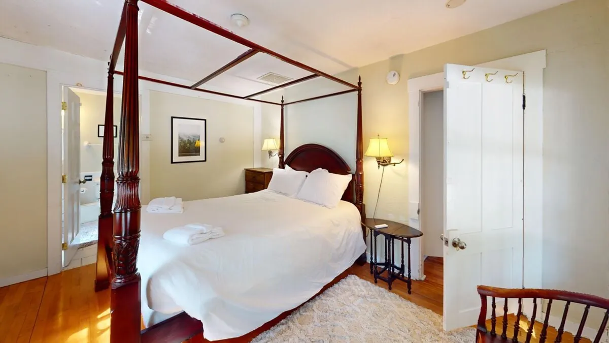 A serene and comfortable bedroom with a four-poster bed, a nightstand and lamp for evening relaxation, framed artwork, and a wooden chair. The space exudes tranquility and is designed for quiet moments of rest, reading, or morning coffee in bed.