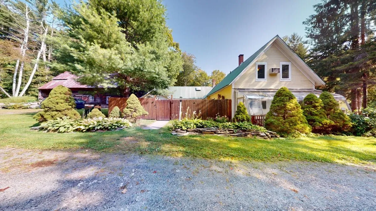 A serene winter-ready exterior view of the yellow farmhouse, highlighted by a wooden fence, manicured shrubs, and a gravel driveway. The setting evokes the charm of Vermont's countryside, with its inviting and peaceful atmosphere ideal for winter getaways.