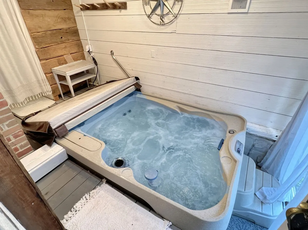 An inviting interior scene featuring a hot tub set against wooden walls, accompanied by a small table and a decorative wheel. The tranquil space is softly lit, exuding warmth and comfort, perfect for relaxing or socializing after outdoor activities.