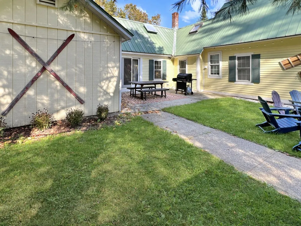 The image features a cheerful yellow house with a green roof, an outdoor dining table, grill, Adirondack chairs, and a decorative cross on the wall. The setting is lively and welcoming, ideal for barbecues, outdoor dining, and relaxed gatherings.