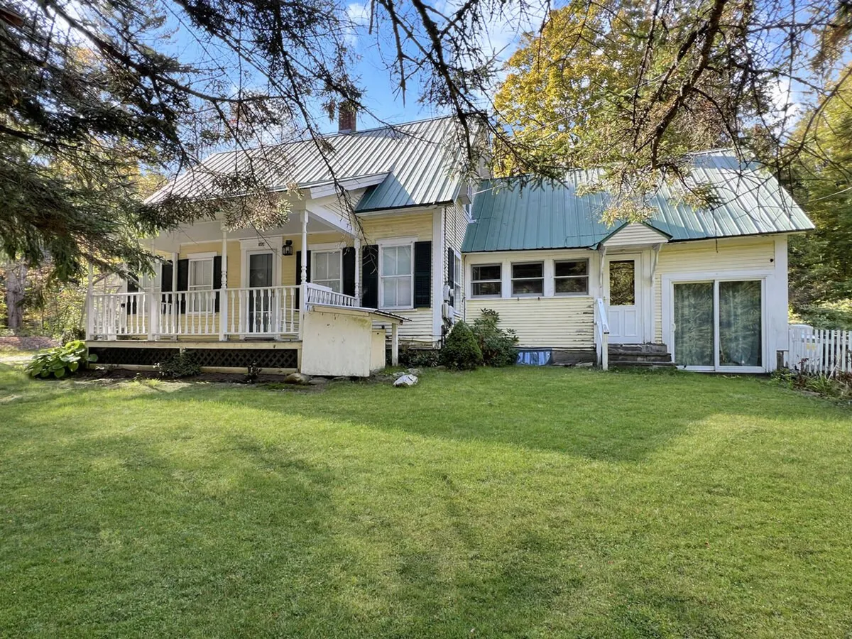 This image features the front porch of the farmhouse with a green roof, surrounded by a lush lawn and mature trees, creating a peaceful and inviting environment. The setting feels secluded and tranquil, ideal for relaxing outdoors or practicing yoga on the grass.