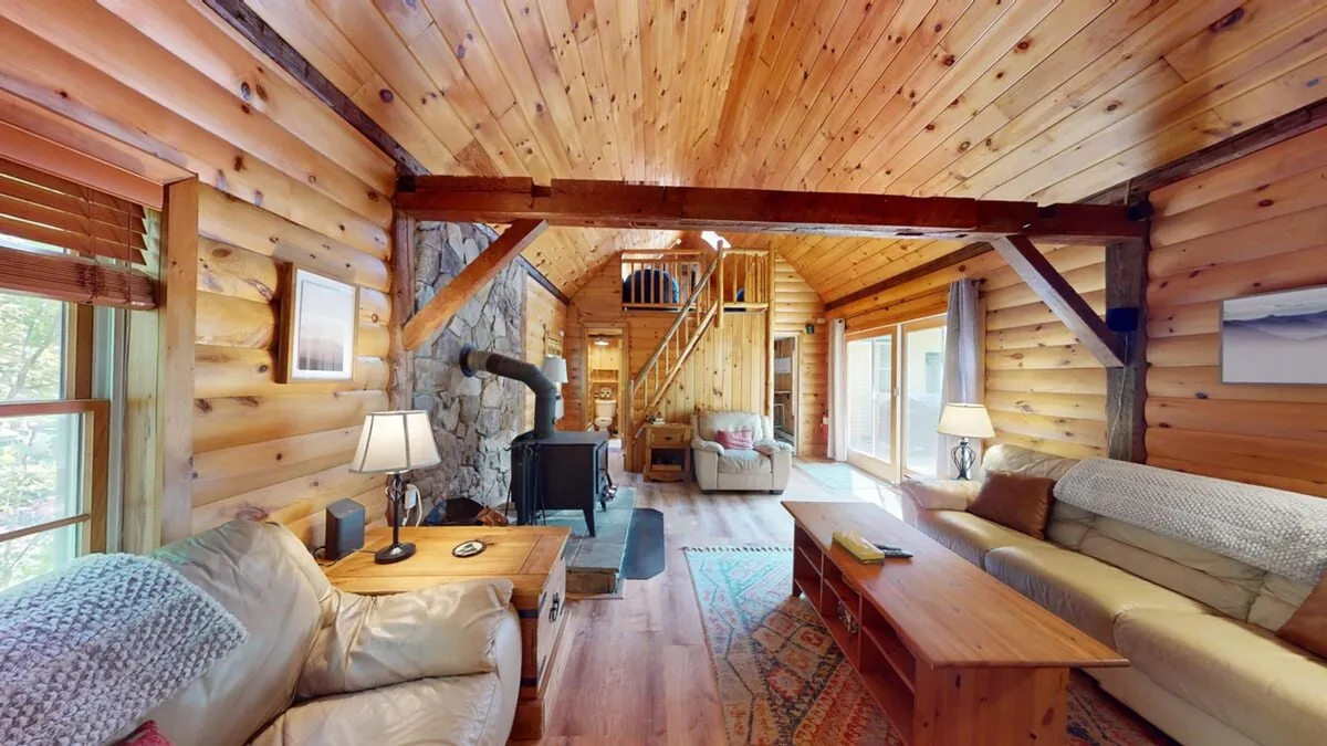 This image features a living room with exposed wooden beams, a grand stone fireplace, large windows with views of nature, comfortable sofas, and a central coffee table. The space feels cozy and inviting, perfect for group gatherings and relaxation.