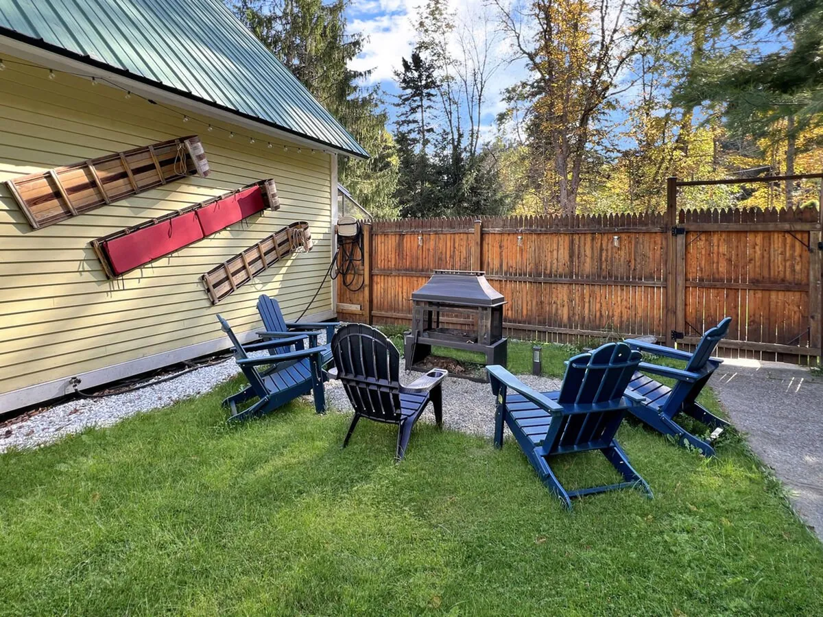 A casual outdoor gathering space featuring blue Adirondack chairs around a fire pit in front of the yellow house. The area is designed for relaxation, socializing, and enjoying the crisp Vermont air after outdoor adventures.