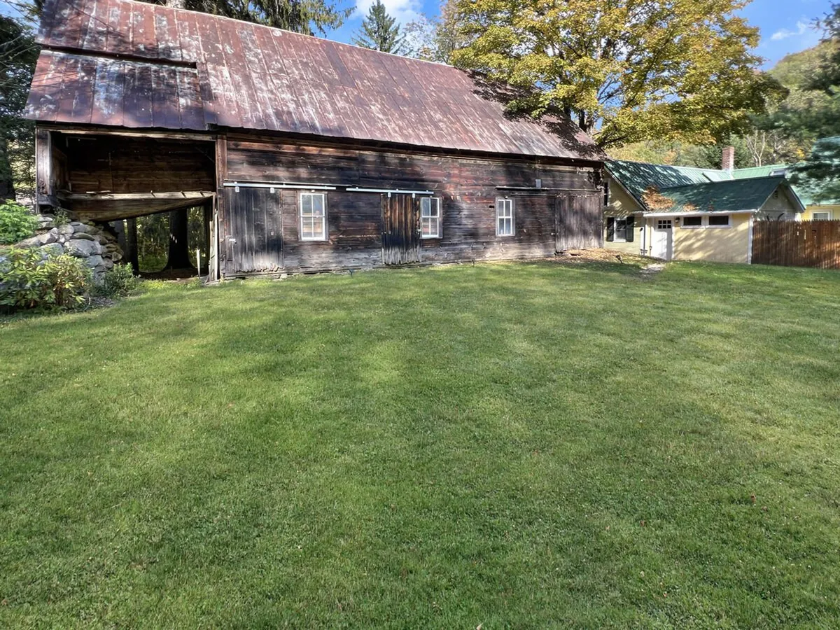 A classic old barn sits on a green lawn, framed by surrounding trees and neighboring structures. The scene evokes nostalgia, history, and a deep sense of place, highlighting the authentic farmstead atmosphere of the property.