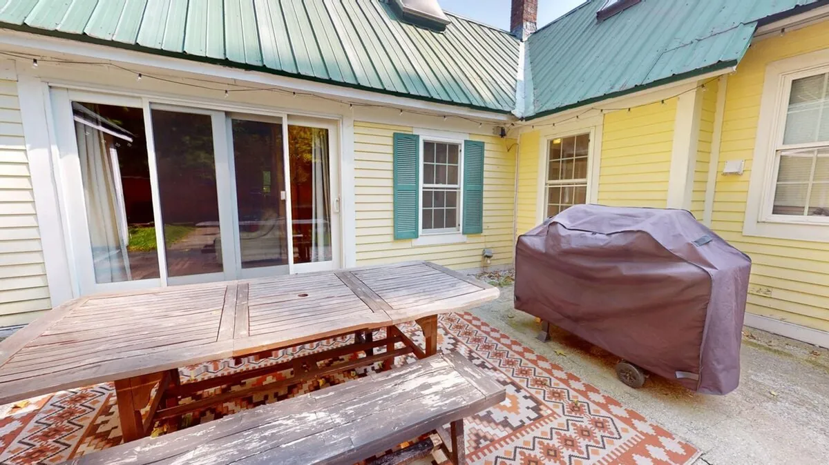An exterior shot of a wooden outdoor dining table with green shutters behind, a grill cover, and a patterned outdoor rug. The space is set up for al fresco dining, barbecues, and relaxed gatherings, with a cozy and casual atmosphere.