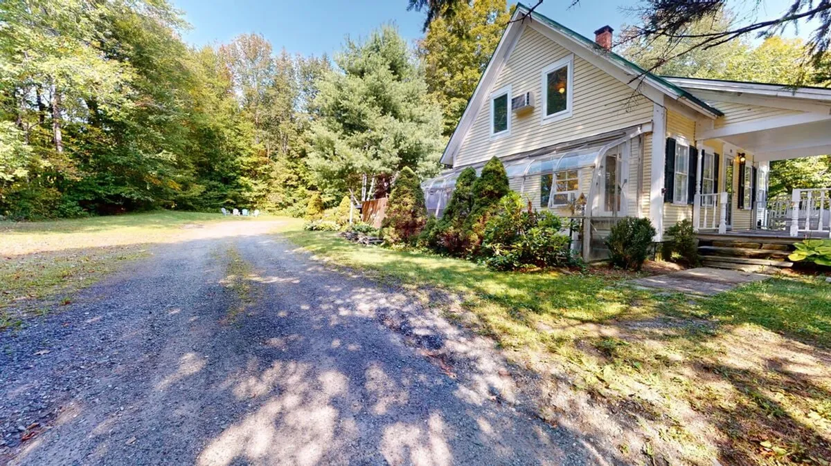 The image depicts the farmhouse from an exterior perspective, surrounded by a landscaped garden, gravel driveway, and mature trees. The warm, welcoming façade invites guests to experience the quintessential Vermont countryside setting, while the garden hints at seasonal outdoor activities and family gatherings.
