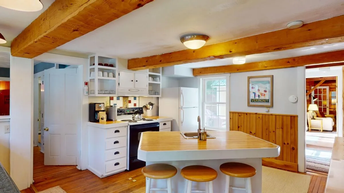 A cozy kitchen scene emphasizing the kitchen island, wooden beams, stove, fridge, and a sunlit window. The space feels intimate but functional, ready for both cooking and socializing, with homey touches evident throughout.