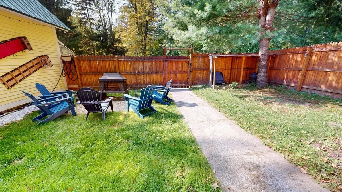 A cluster of Adirondack chairs along a stone patio pathway with a grill nearby, set against a backdrop of greenery. The setup encourages relaxing, sunset views, and small group gatherings in a laid-back outdoor space.